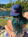 a woman in a Zilker Belts Freedom Rope Hat sitting on a blanket next to a lake.