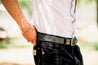 Man wearing a white t-shirt and black jeans, tightening a Zilker Belts Maverick black leather belt with a metal buckle.