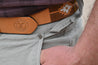Close-up of a person wearing gray pants and a brown belt with a stamped leather tag from the Zilker Belts "Texas Wildlife Association (PREORDER).