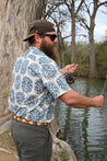 A bearded man wearing a Zilker Belts Texas Wildlife Association patterned shirt and a hat fly fishing by a river, holding a fishing rod with the line extended over the water.