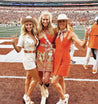Three women in orange hats from the Texas Exes organization posing on a football field in the Zilker Belts area.