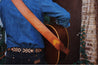 A man is holding a Zilker Belts Vagabond hand-stitched acoustic guitar in front of a brick wall.