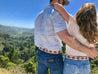 a man and woman hugging while standing on a hill overlooking a valley, wearing Zilker Belts' ATX Light.