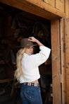 Girl wearing a light brown leather belt with black and white stitching. She styles it with dark blue jeans, a white top, and a cowboy hat.