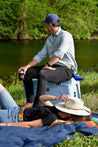 A man and woman sitting on a Rambler blanket next to a river.