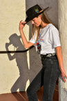 A woman wearing a Midnight Rider cowboy hat and jeans leaning against a wall, all accessorized with Zilker Belts.