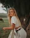 A blonde woman in a white crop top and white pants leaning against a horse wearing Zilker Belts' Reveille.