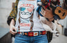 A woman wearing a Willie t-shirt is holding a dog and a ukulele.